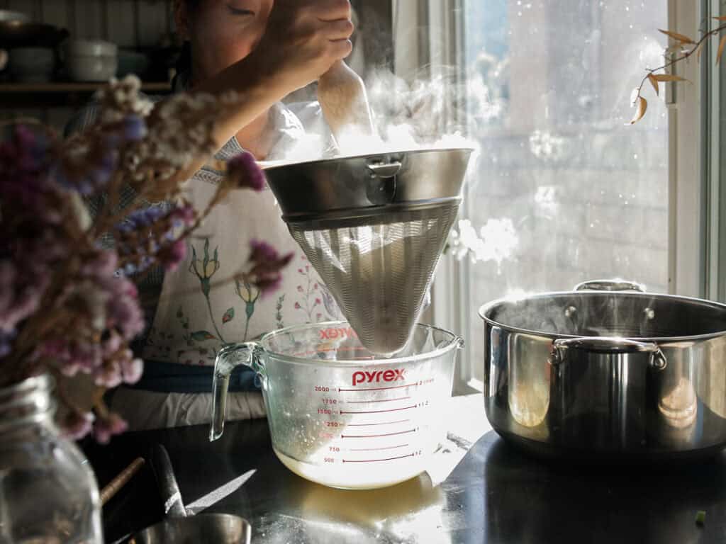 A person strains hot broth through a metal sieve into a glass measuring cup. Sunlight streams through a nearby window, illuminating steam rising from the pot. A bouquet of flowers sits in the foreground.